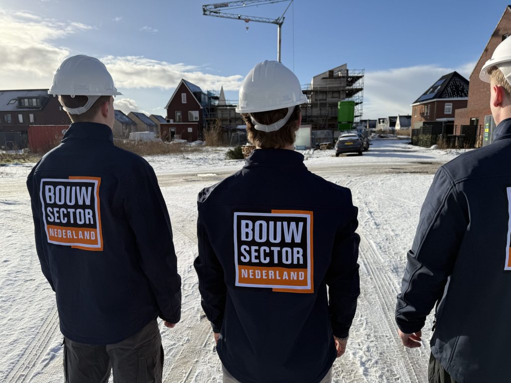Drie vakmensen van Bouwsector Nederland met witte helmen en jassen met logo kijken naar nieuwbouwwoningen in aanbouw op een besneeuwde bouwplaats